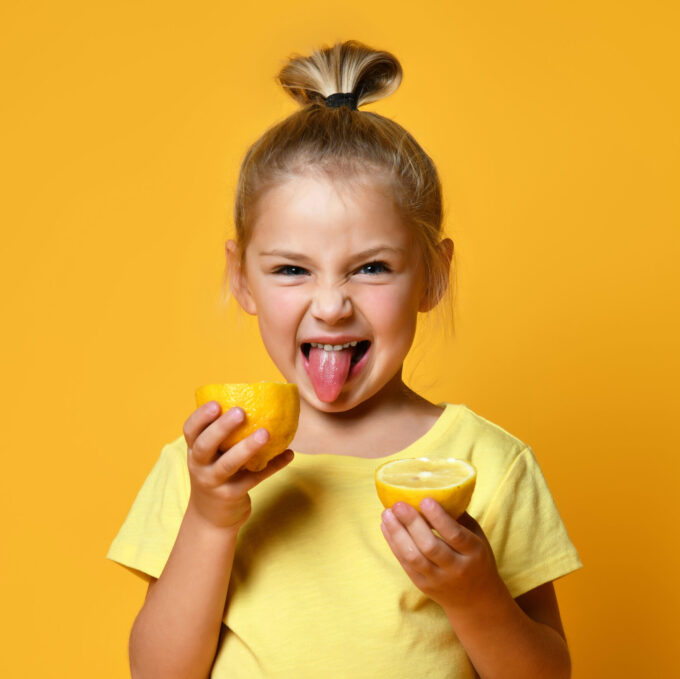 Little smiling cute blond girl in yellow t-shirt holding halves of fresh sour ripe lemon fruit and showing tongue over yellow background. Healthy lifestyle and clean eating concept